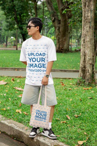 Oversized T-Shirt Mockup of a Smiling Man Carrying a Tote Bag in a Park