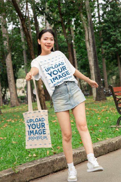 Oversized T-Shirt Mockup of a Woman Showing Her Tote Bag in a Park